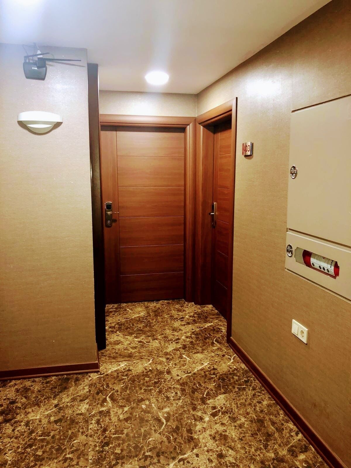 Un couloir étroit avec des portes en bois, un sol en marbre et un tableau électrique fixé au mur.