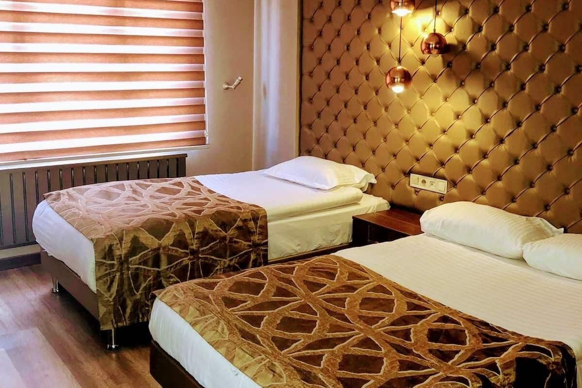 A hotel room with two beds, a tufted headboard, a window with blinds, and a hanging light fixture.