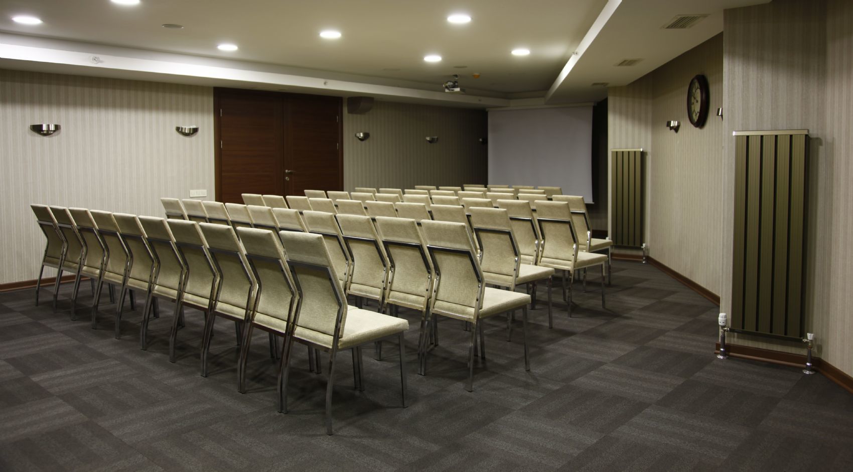 Een vergaderzaal met rijen stoelen, een projectiescherm en een klok aan de muur.