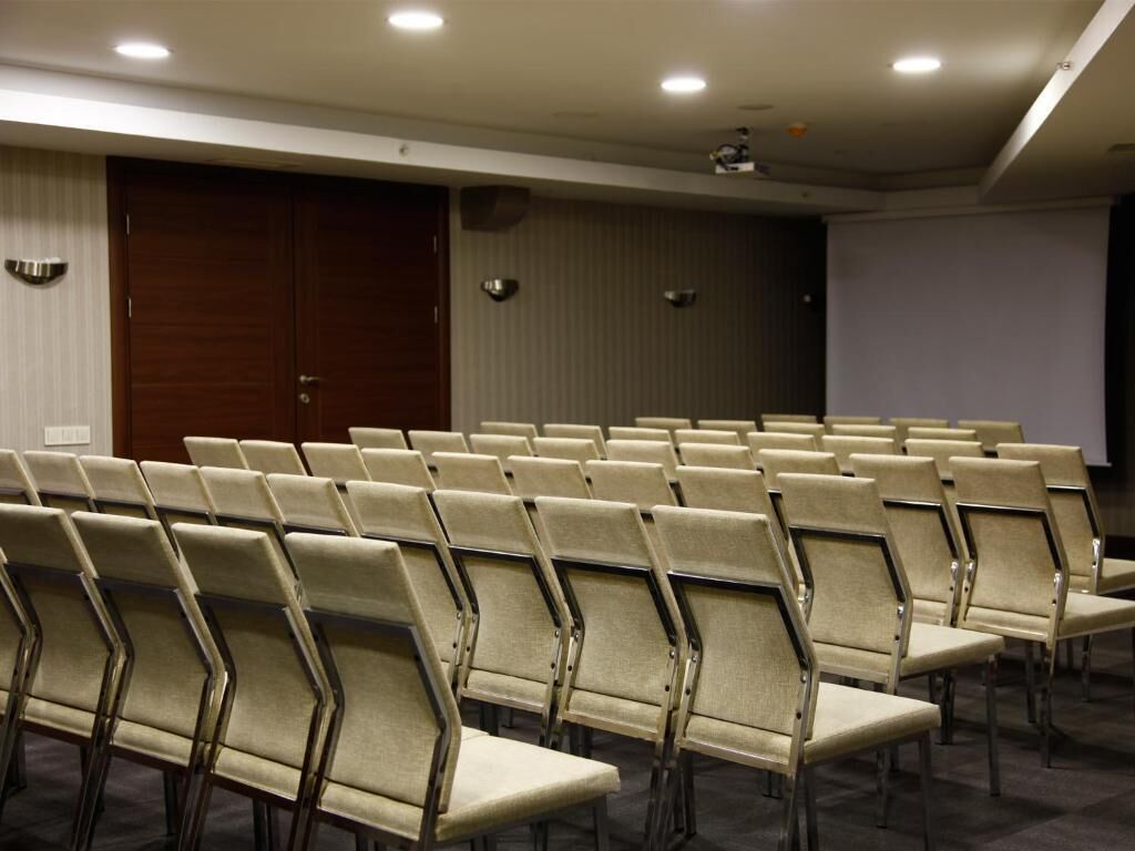 Une salle de conférence avec des rangées de chaises beiges, un écran de projection et une porte fermée.