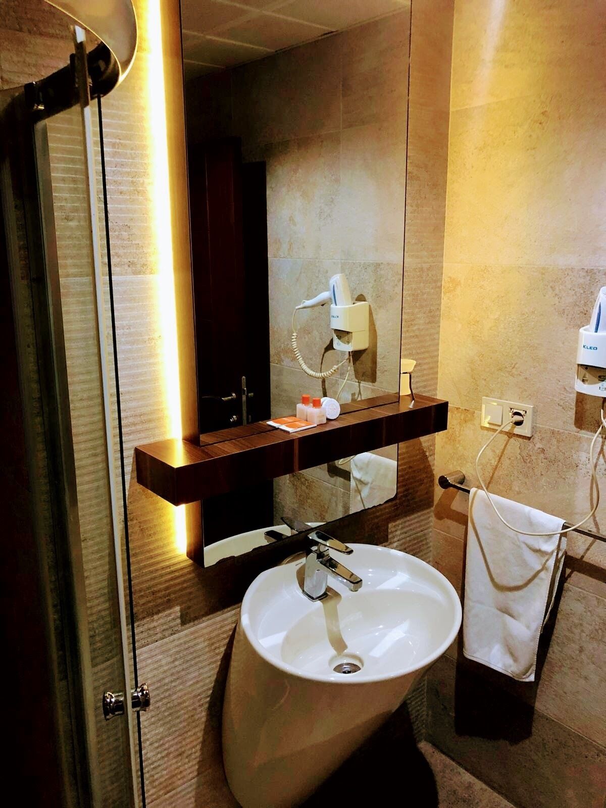 A modern bathroom with a glass shower enclosure, a white sink, a towel rack, and a hairdryer mounted on the wall.