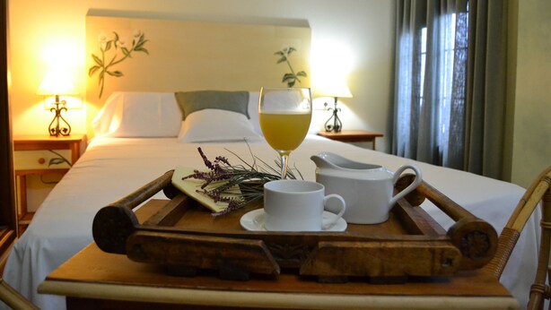 A neatly made bed with white linens, a bedside table with a lamp, a tray with a glass of juice, a cup, and a pitcher. - Hostal Toribio (Galaroza)