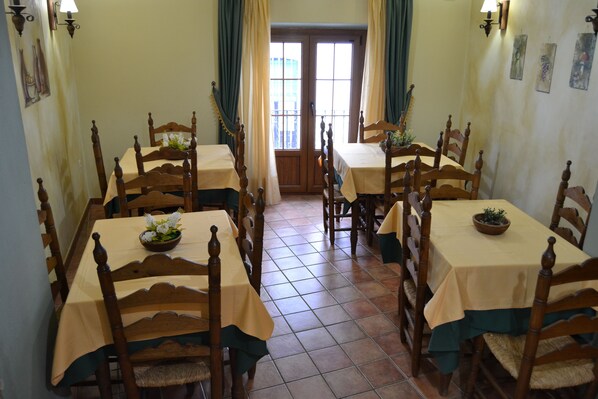 Un comedor con mesas y sillas de madera, suelo de baldosas y vistas a través de una puerta con cortinas.