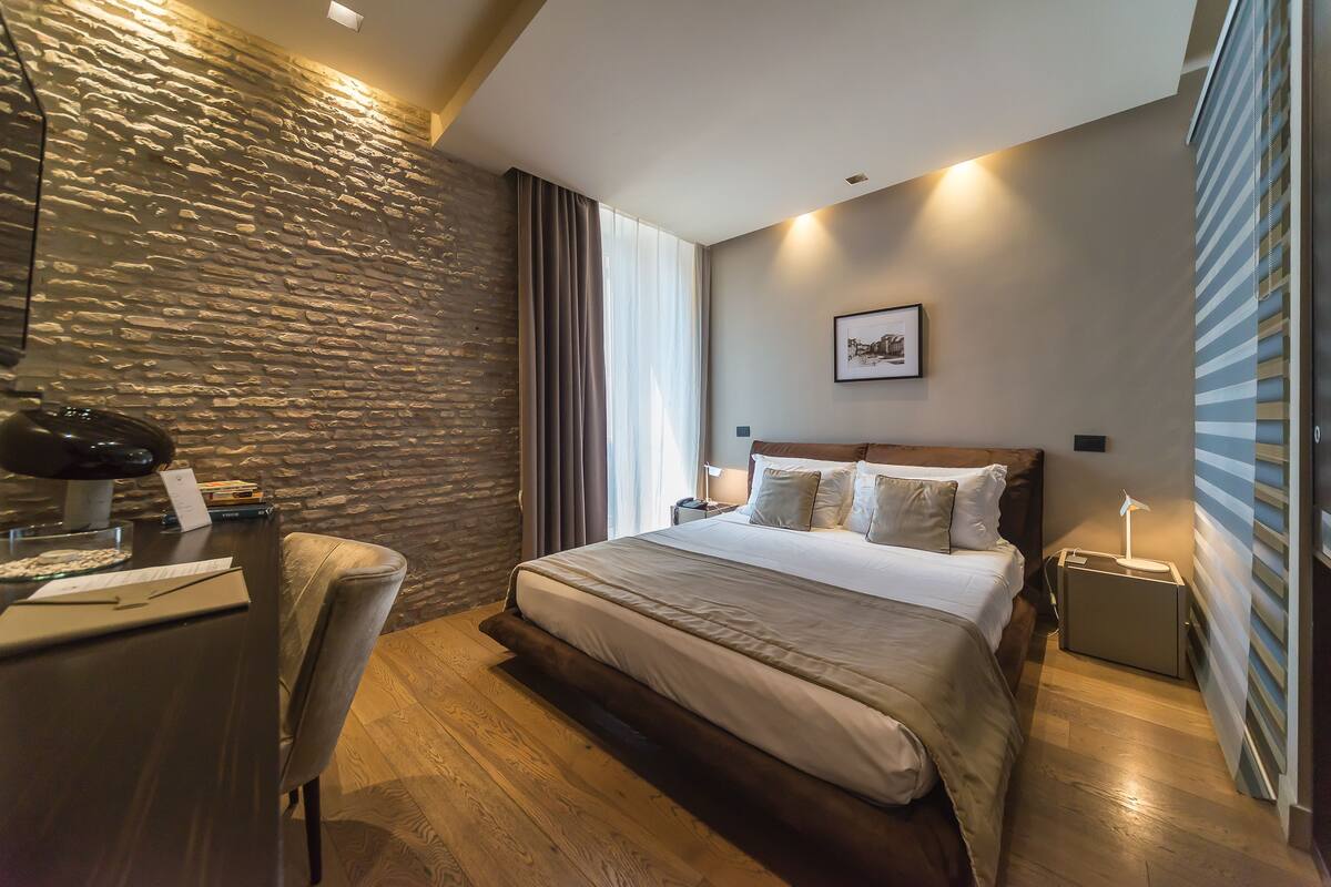 A modern hotel room with a stone accent wall, a large bed, a desk with a chair, and a bedside table with a lamp.