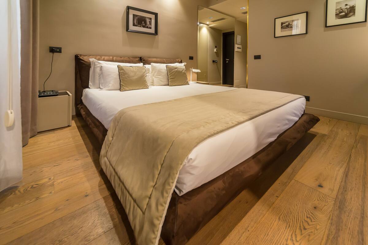 A bedroom with a large bed, wooden flooring, and a nightstand with a lamp.