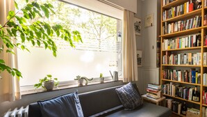 A cozy reading nook with a sofa, a bookshelf, and a window with plants.