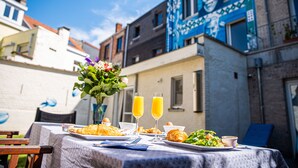 An outdoor dining table with breakfast items and drinks, set against a backdrop of colorful buildings.
