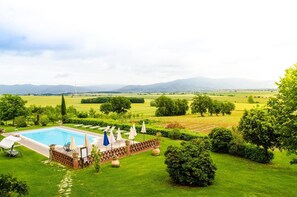 Une piscine entourée de chaises longues et de parasols, bordée de pelouses et d’arbres bien entretenus, et offrant une vue sur les montagnes lointaines.