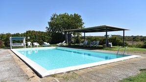 Una piscina rectangular con un área de terraza cubierta, rodeada de sillas de descanso y un pabellón.