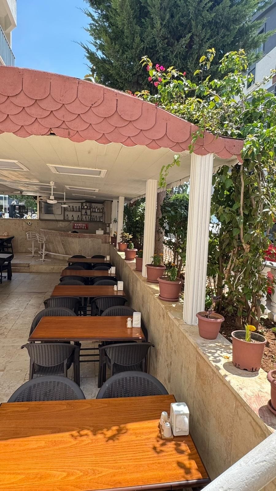 Un espacio al aire libre con mesas y sillas, una sección cubierta con techo de baldosas y plantas en macetas.