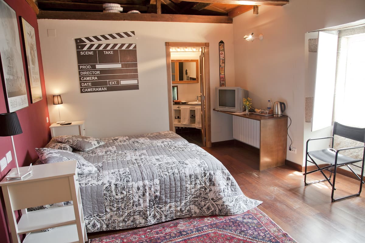 A bedroom with a bed, a desk, a chair, a television, and a bathroom visible through an open door.