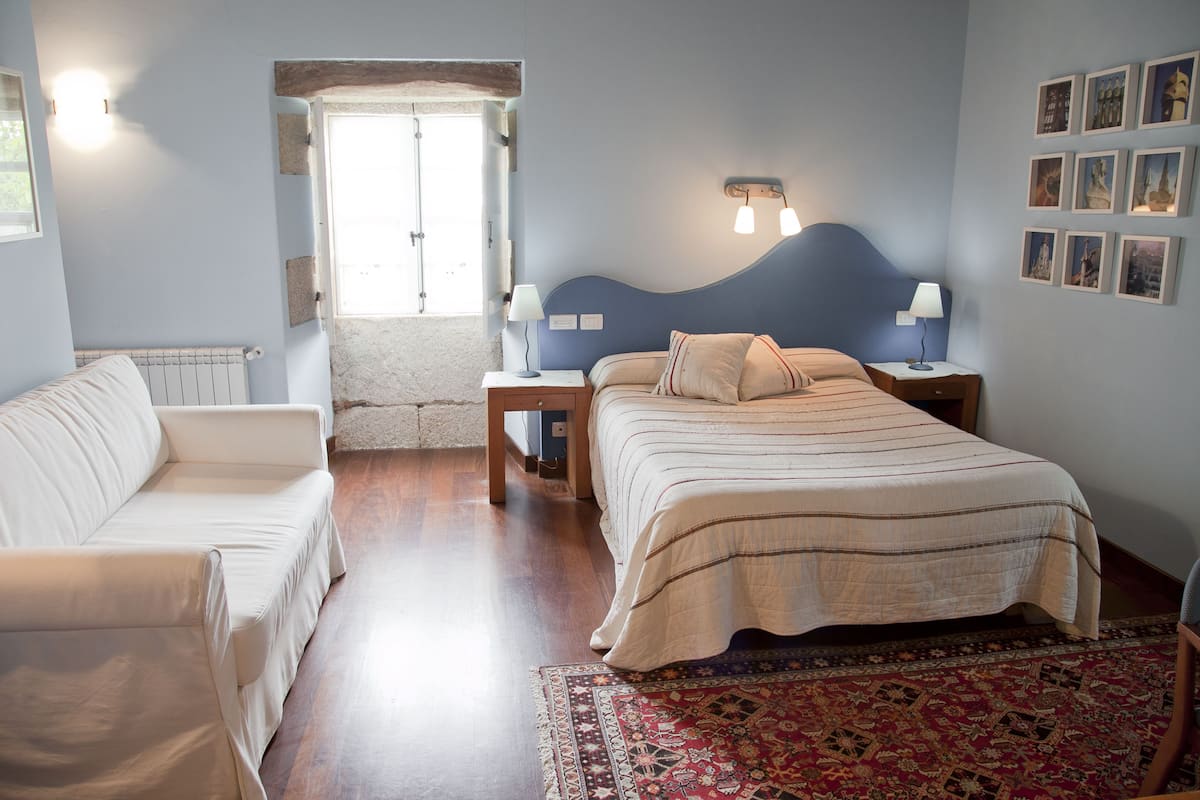 A bedroom with a bed, a sofa, a nightstand, a lamp, a radiator, and a window.
