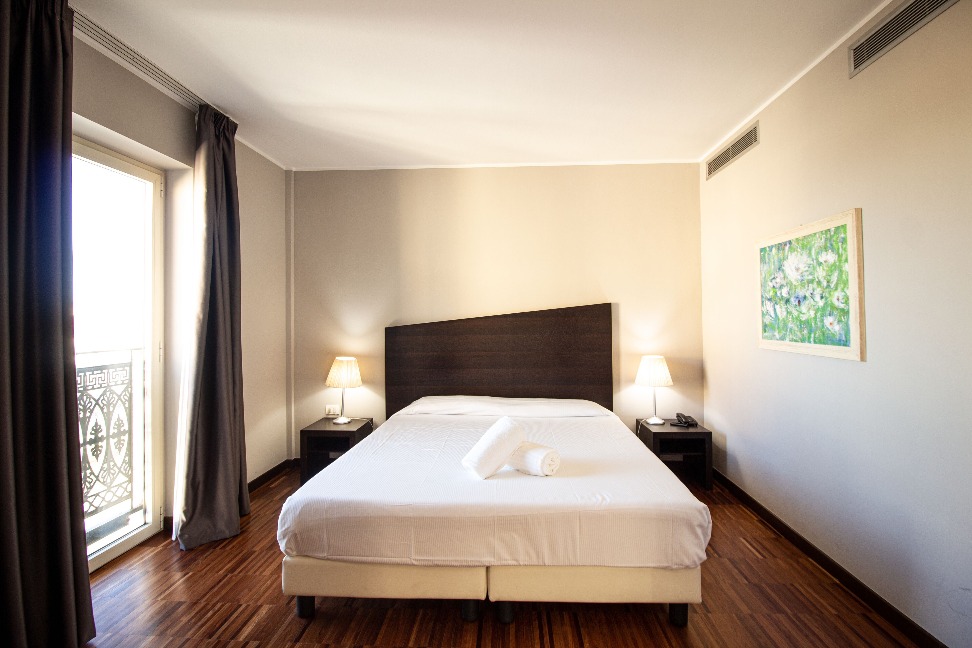 A hotel room with a large bed, two bedside tables with lamps, a painting on the wall, and a view through a window with curtains.
