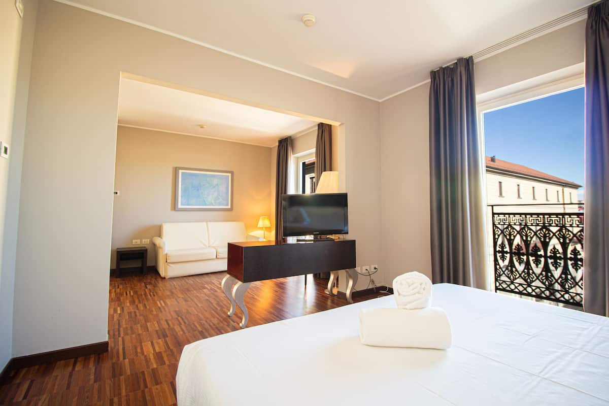 A hotel room with a bed, a TV, a desk, a chair, a sofa, and a balcony with a view of buildings.