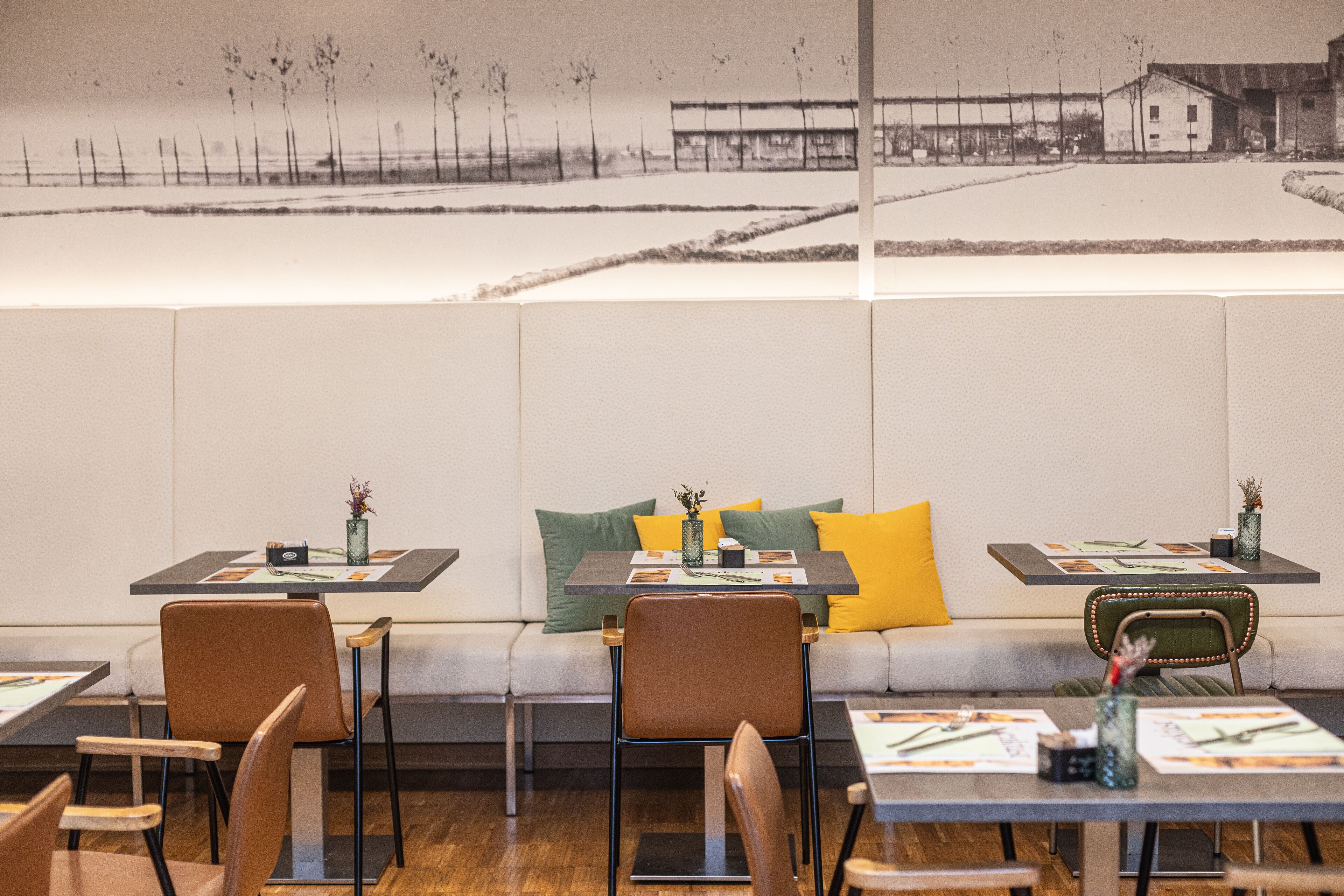 A restaurant with tables set for dining, a sofa with cushions, and a wall-mounted artwork depicting a rural scene.