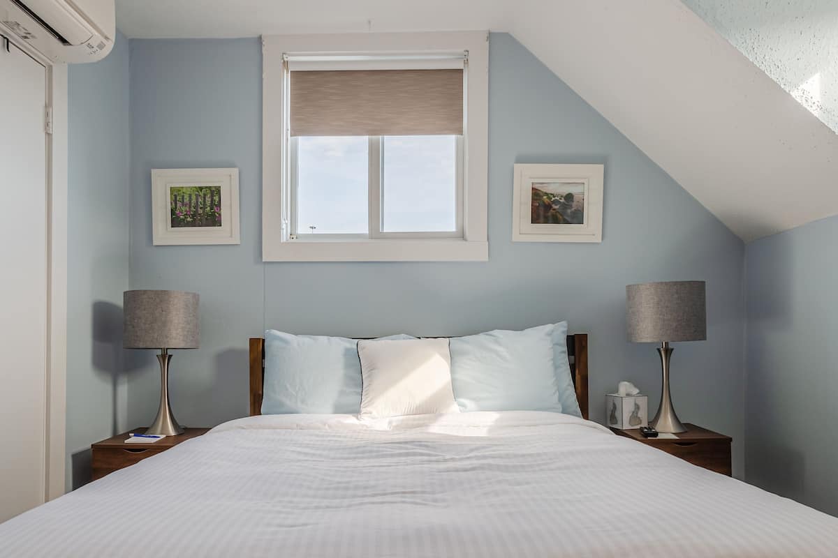 A bedroom with a bed, two bedside tables with lamps, a window with blinds, and two framed pictures on the wall.