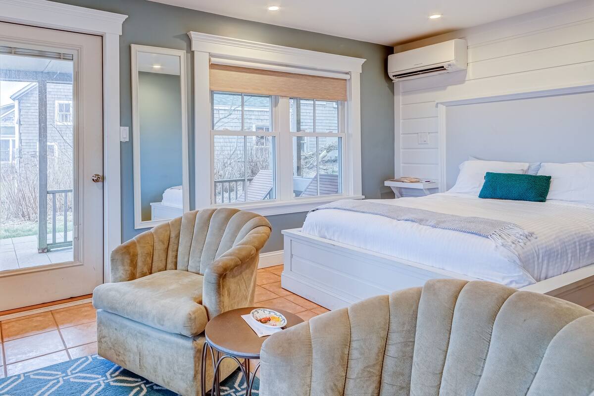 A bedroom with a bed, two armchairs, a small table, and a window with a view of a house and trees.