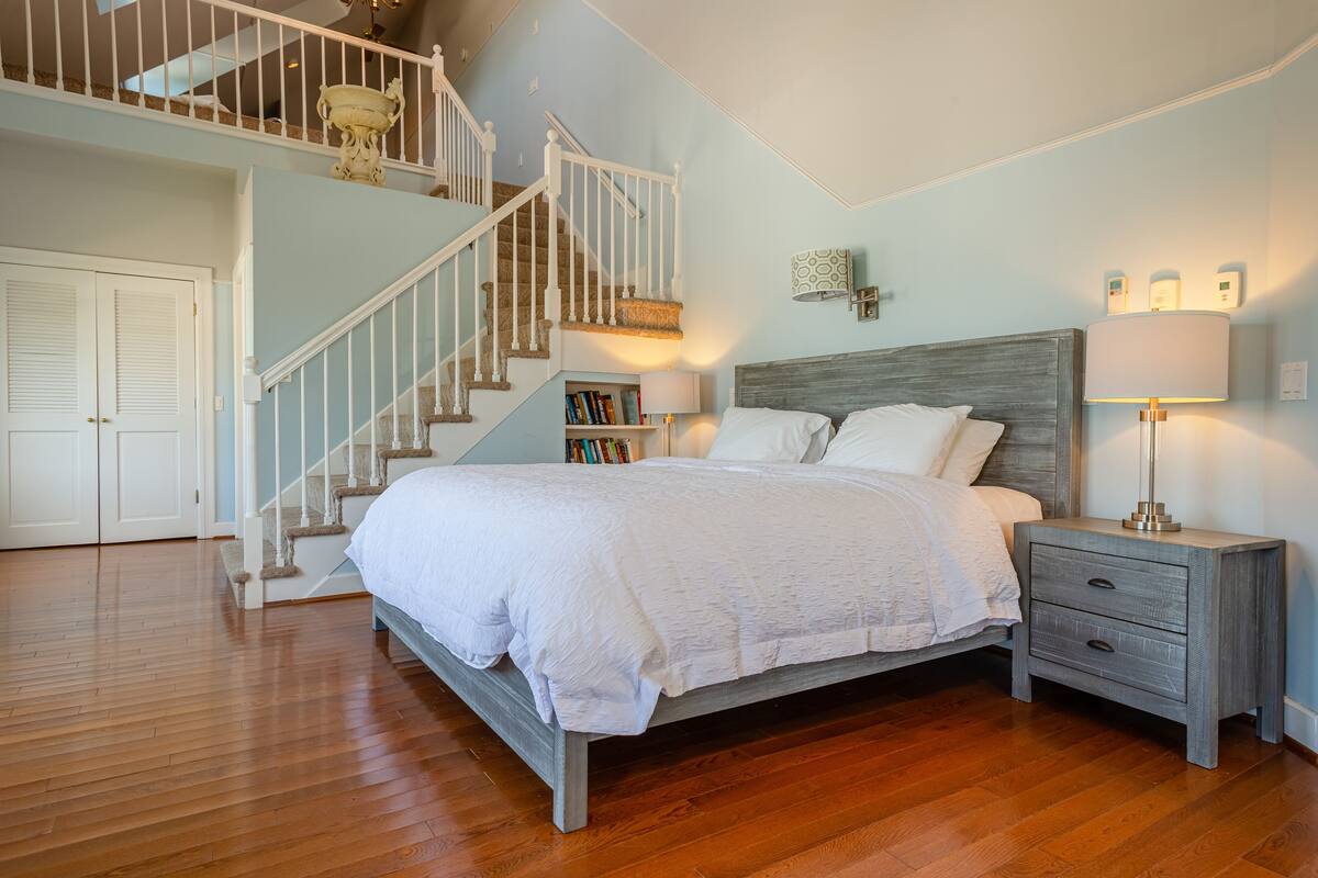 A bedroom with a wooden floor, a bed with white bedding, a nightstand with a lamp, a bookshelf, and a staircase with a white railing.