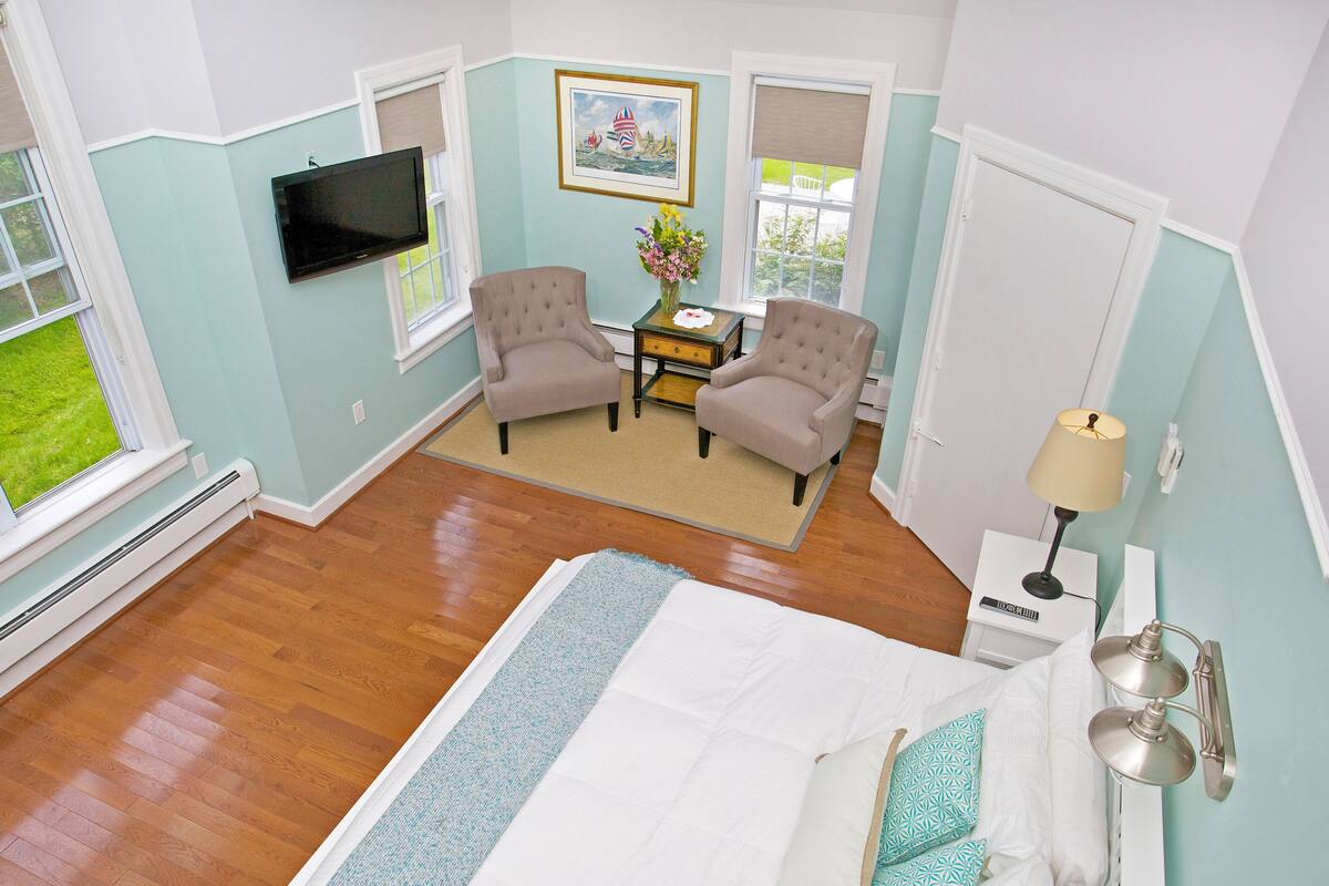 A hotel room with a bed, two armchairs, a small table with a vase, a TV, and a window with a view of greenery.