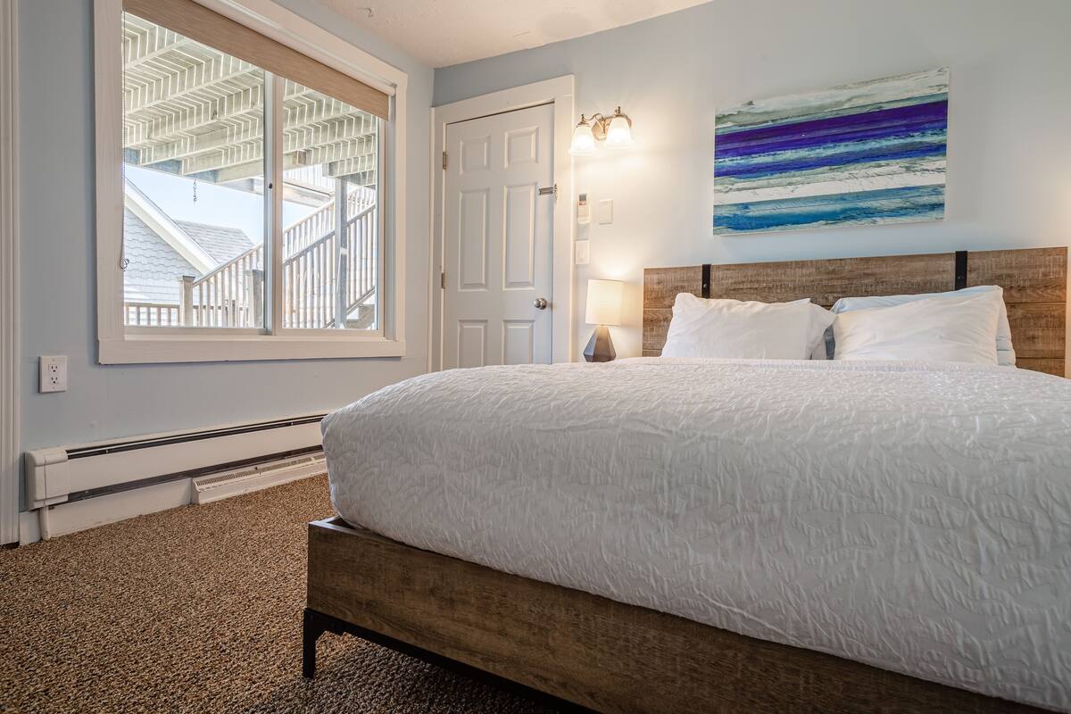 A bedroom with a large bed, a nightstand, a wall-mounted lamp, and a window with a view of a staircase.