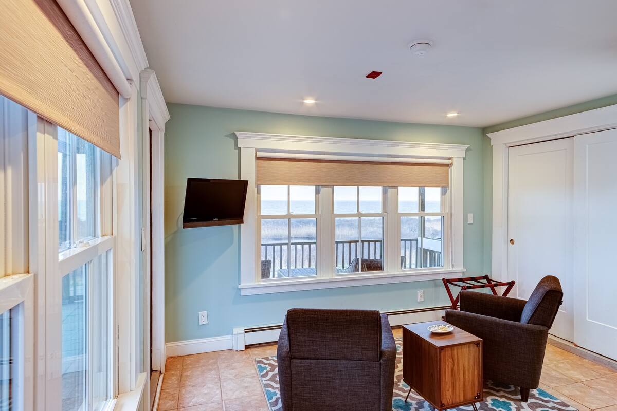 A room with a large window, a flat-screen TV mounted on the wall, two chairs, and a small table.