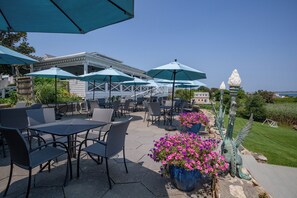 An outdoor patio with tables and chairs, covered by blue umbrellas, and surrounded by potted plants and decorative statues.