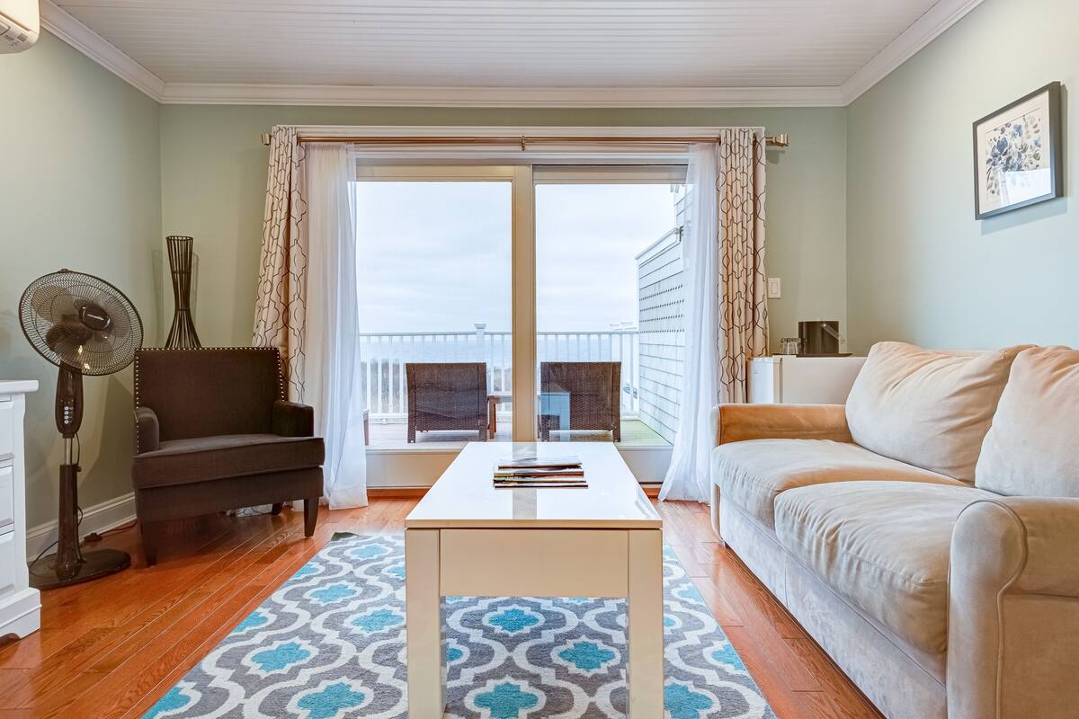 A living room with a sofa, a coffee table, a fan, and a view through a glass door to an outdoor balcony.
