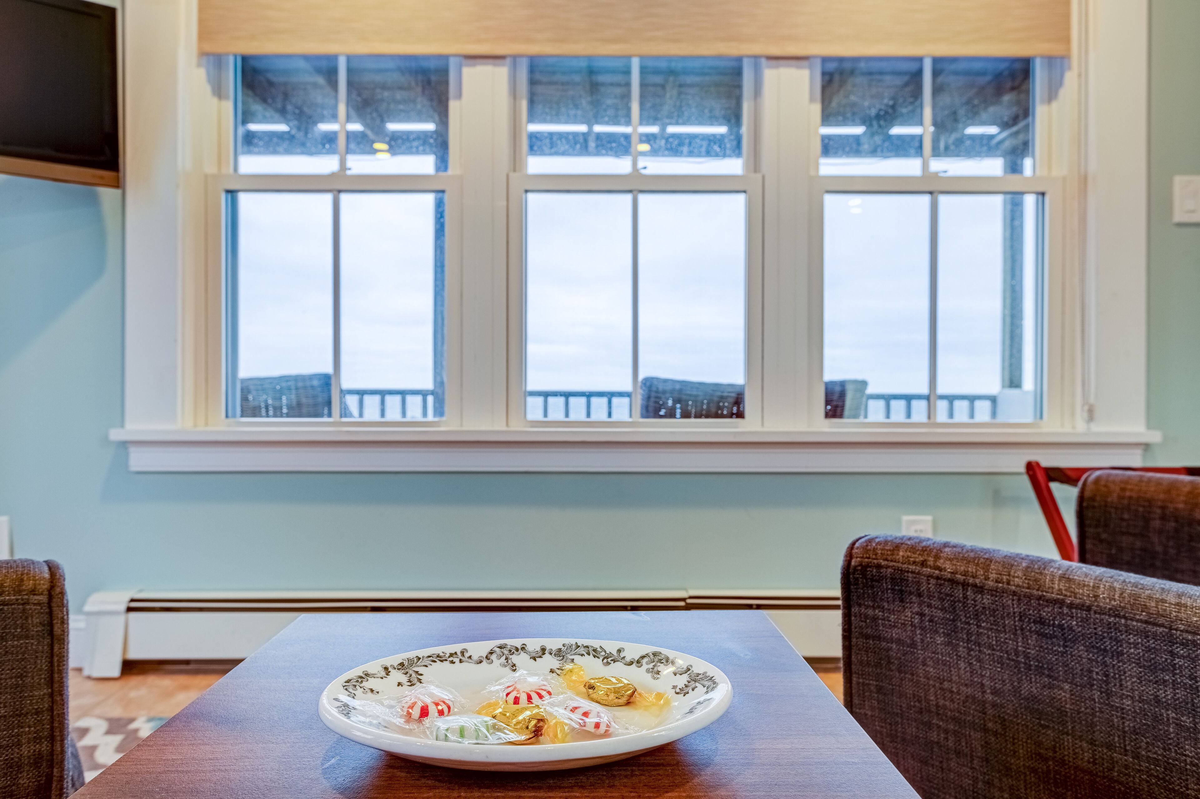 Un comedor con una mesa dispuesta para dos personas, una ventana que da a un balcón y un plato con caramelos variados sobre la mesa.