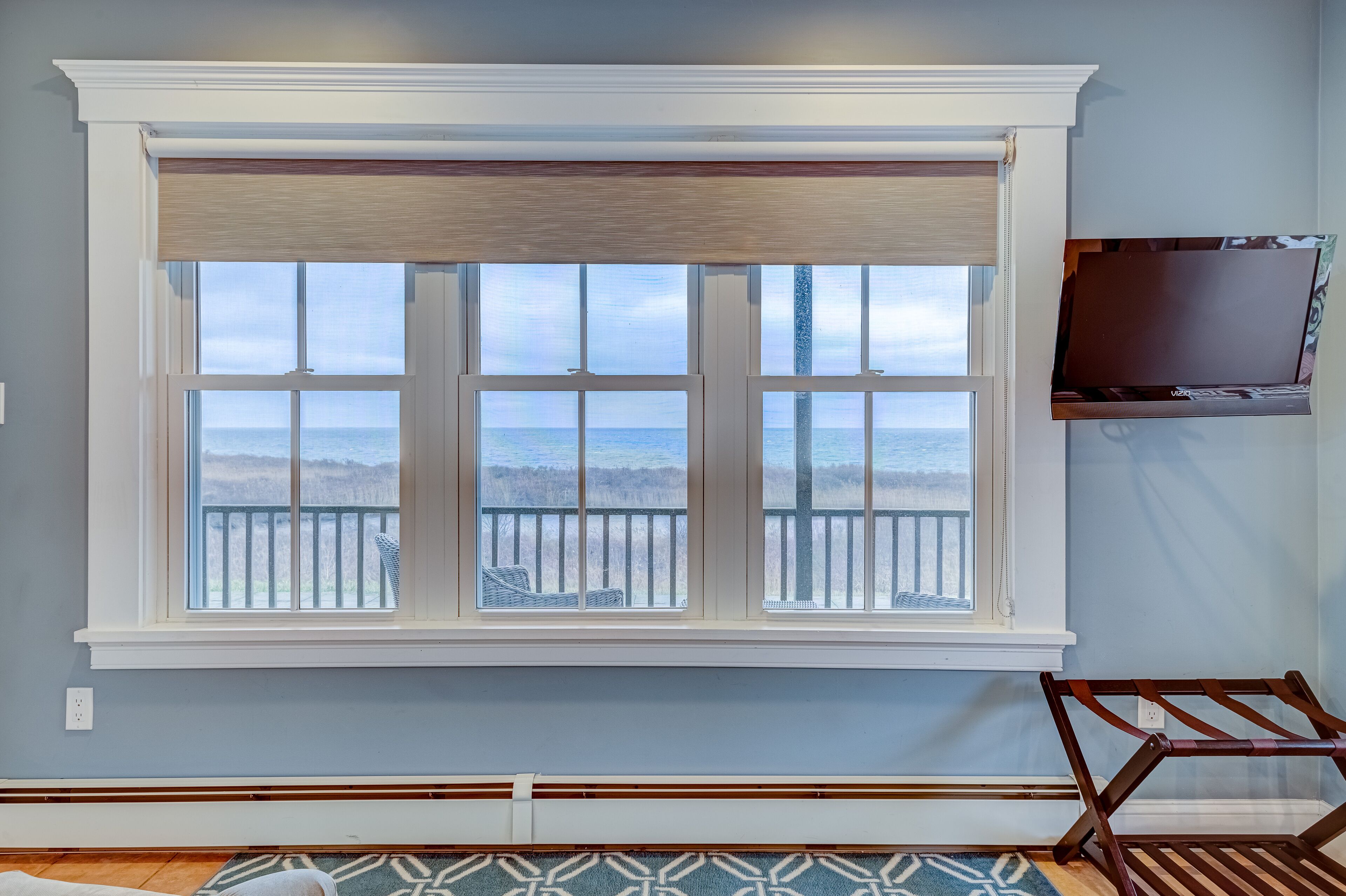 A room with a large window overlooking a beach, a television mounted on the wall, and a patterned rug on the floor.