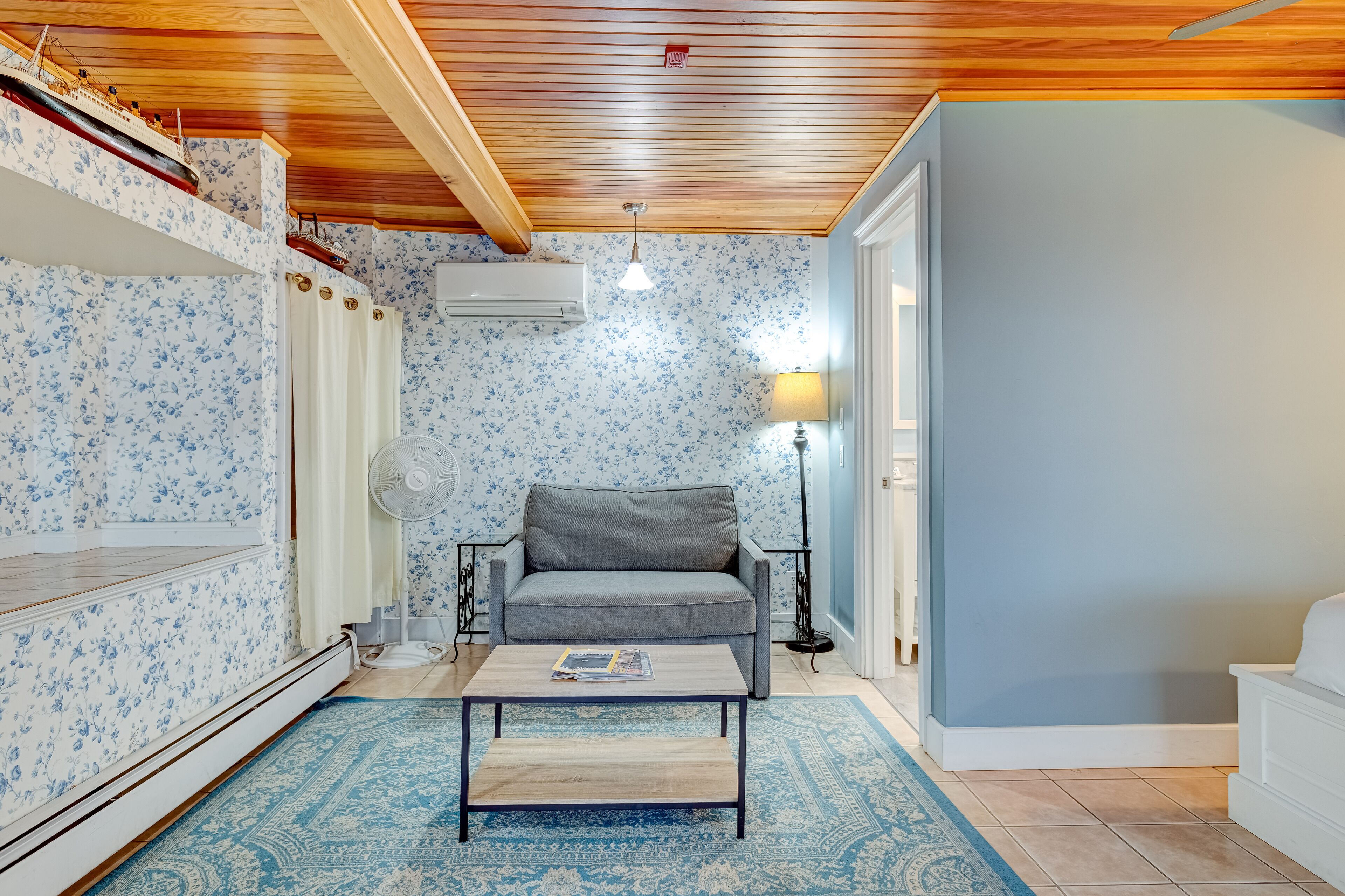Una habitación con techo de madera, papel tapiz floral, un sofá gris, una mesita con una revista, una lámpara de pie y un ventilador de pie.