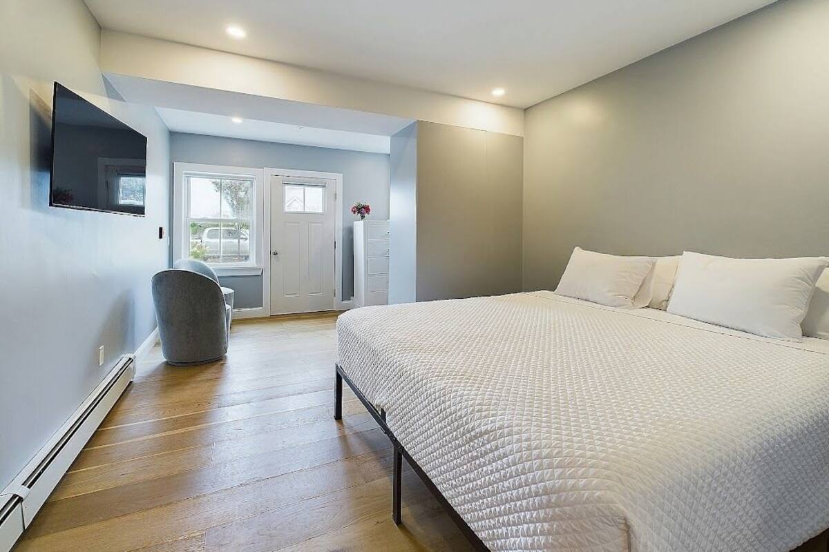 Een slaapkamer met een groot bed, een stoel, een aan de muur bevestigde televisie en een deur die leidt naar een balkon.
