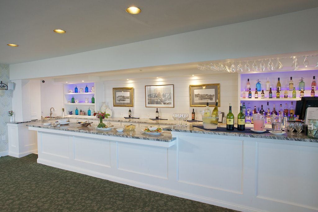 A bar area with a marble countertop, shelves with bottles, framed pictures, and a variety of glasses.