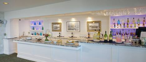 A bar area with a marble countertop, shelves with bottles, framed pictures, and a variety of glasses.