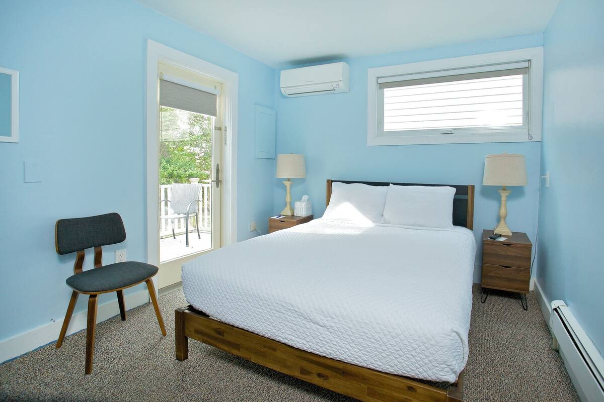 A bedroom with a wooden bed, two bedside tables with lamps, a chair, and a window with blinds.