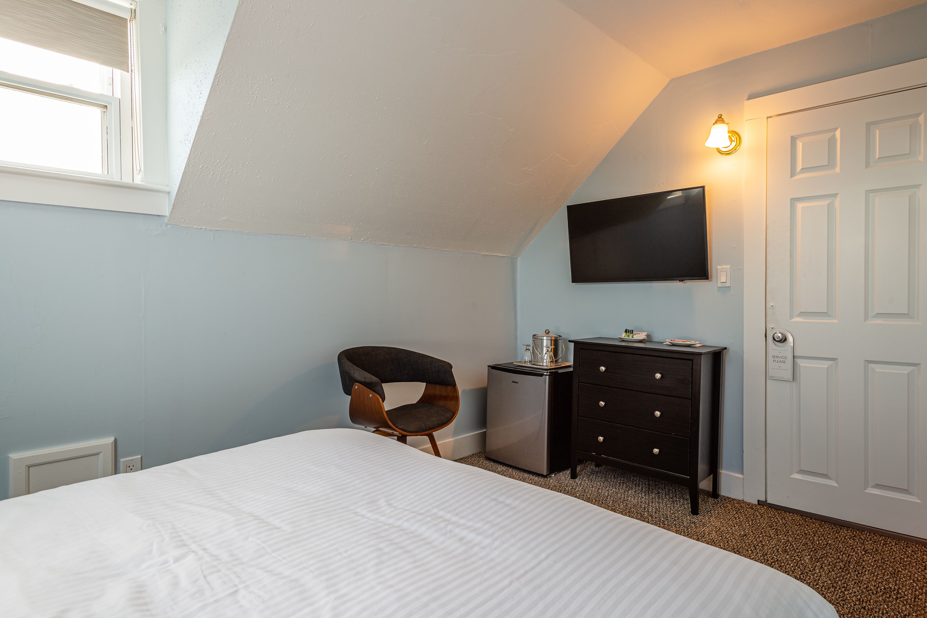 Un dormitorio con cama, escritorio con computadora, silla, televisor montado en la pared y una puerta.