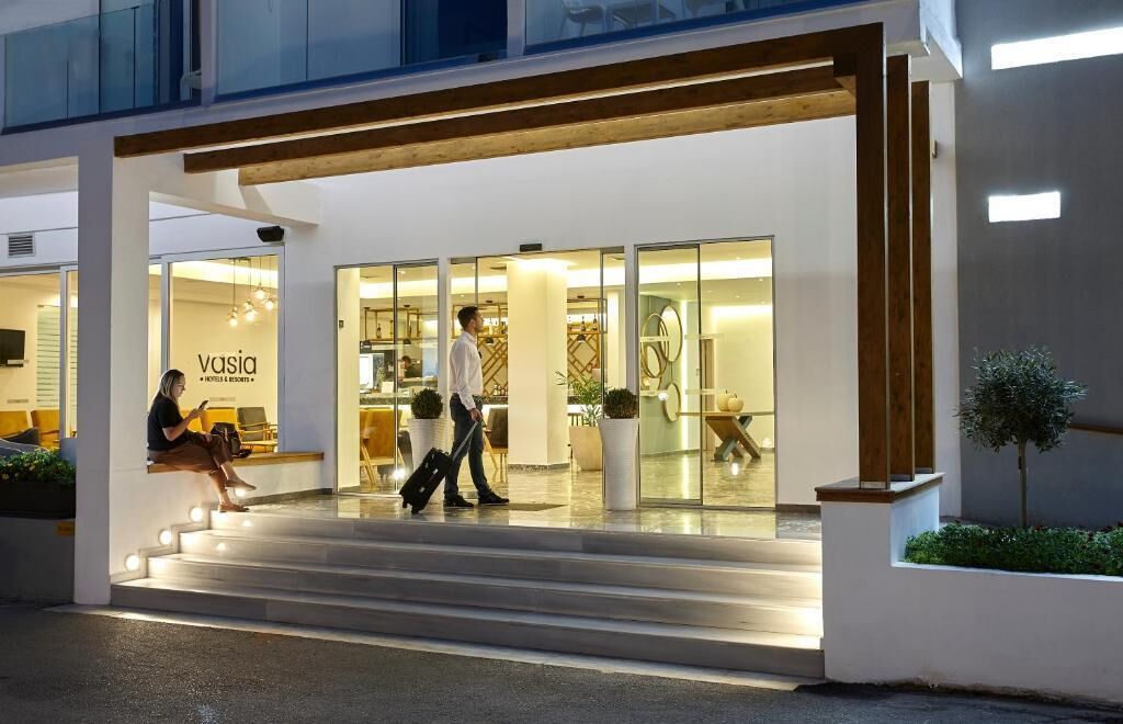 A modern hotel entrance with a glass facade, a reception desk, and a staircase leading to the lobby.