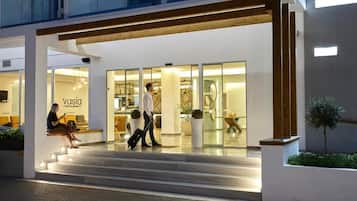 A modern hotel entrance with a glass facade, a reception desk, and a staircase leading to the lobby.