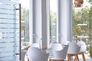 Un espace repas avec des chaises blanches et une table, de grandes fenêtres et une vue sur la verdure à l’extérieur.