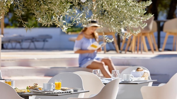 Un espace de restauration en plein air avec des chaises et des tables blanches, une personne qui lit un livre et une table avec des aliments pour le petit-déjeuner.
