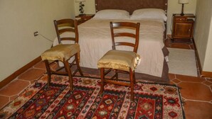 Un dormitorio con una cama, dos sillas de madera, una mesita de noche con lámpara y un tapete estampado.