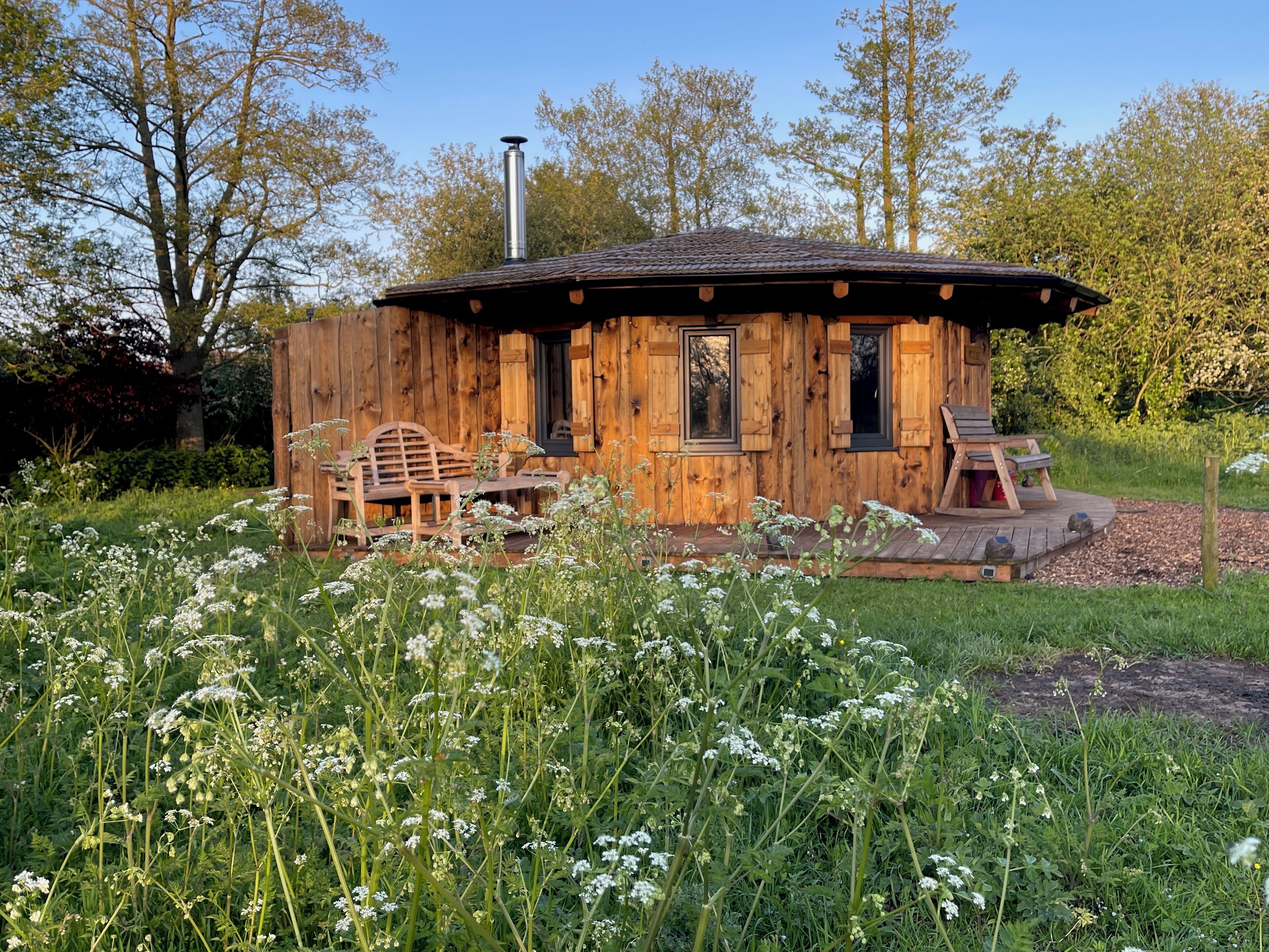 Eine rustikale Holzhütte mit Schornstein, einer Veranda mit Sitzgelegenheiten und einem Garten mit Wildblumen.