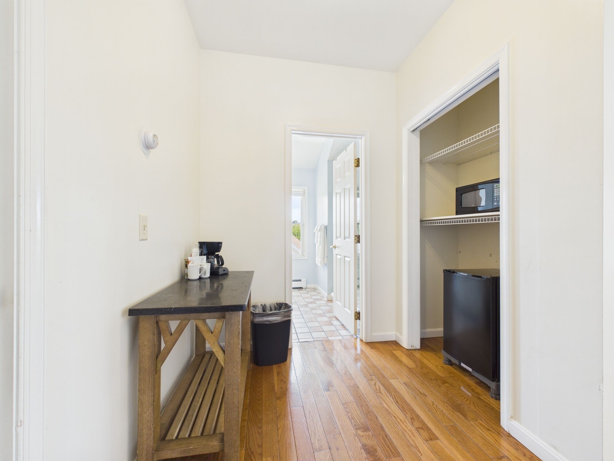 Un pasillo estrecho con suelo de madera, una pequeña mesa con cafetera, un cesto de basura y un refrigerador.