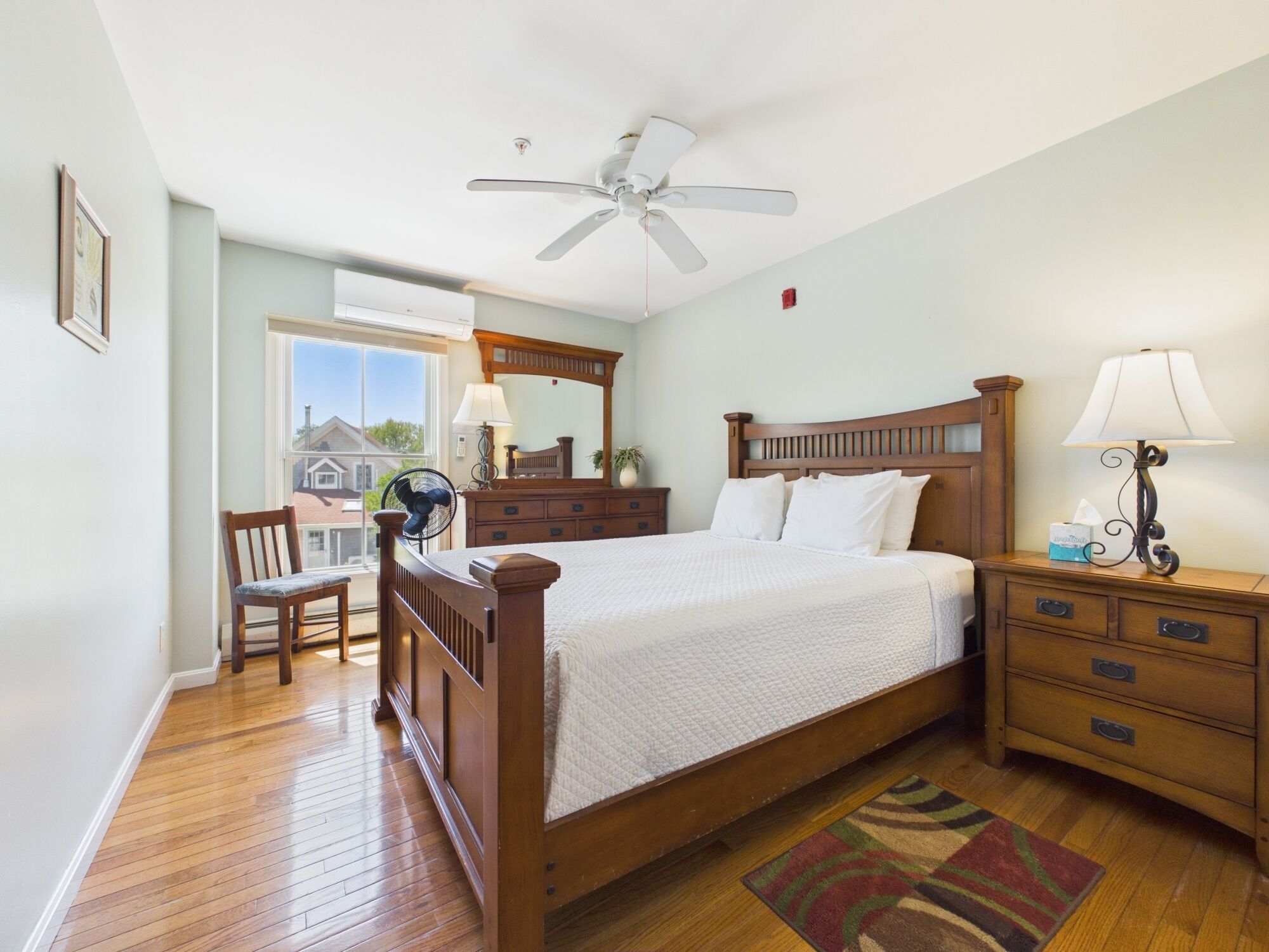 A bedroom with a wooden bed, a nightstand, a chair, a ceiling fan, and a window with a view of houses.