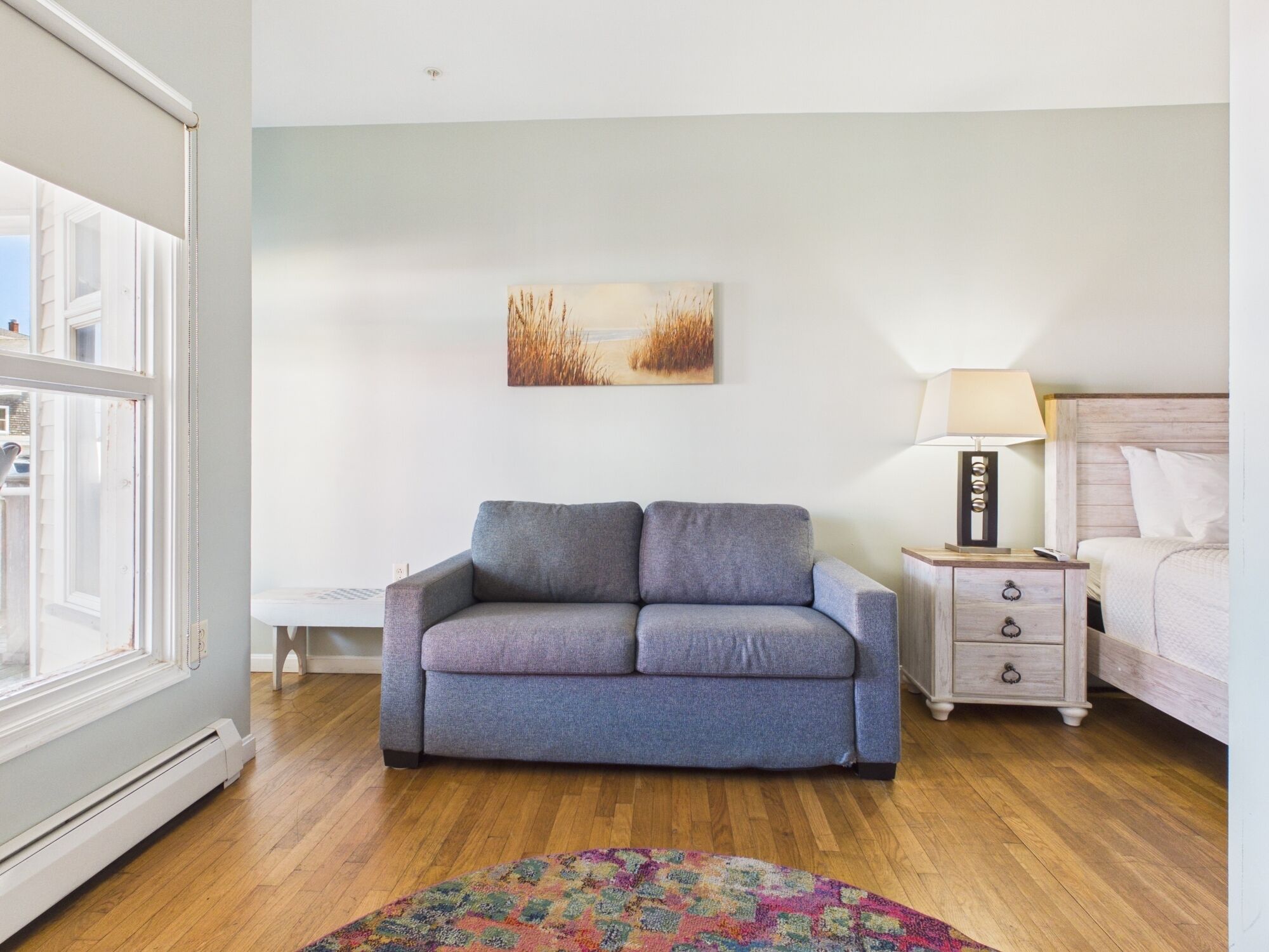 A room with a grey sofa, a small bedside table with a lamp, a bed with white bedding, a colorful rug, and a painting of tall grass on the wall.