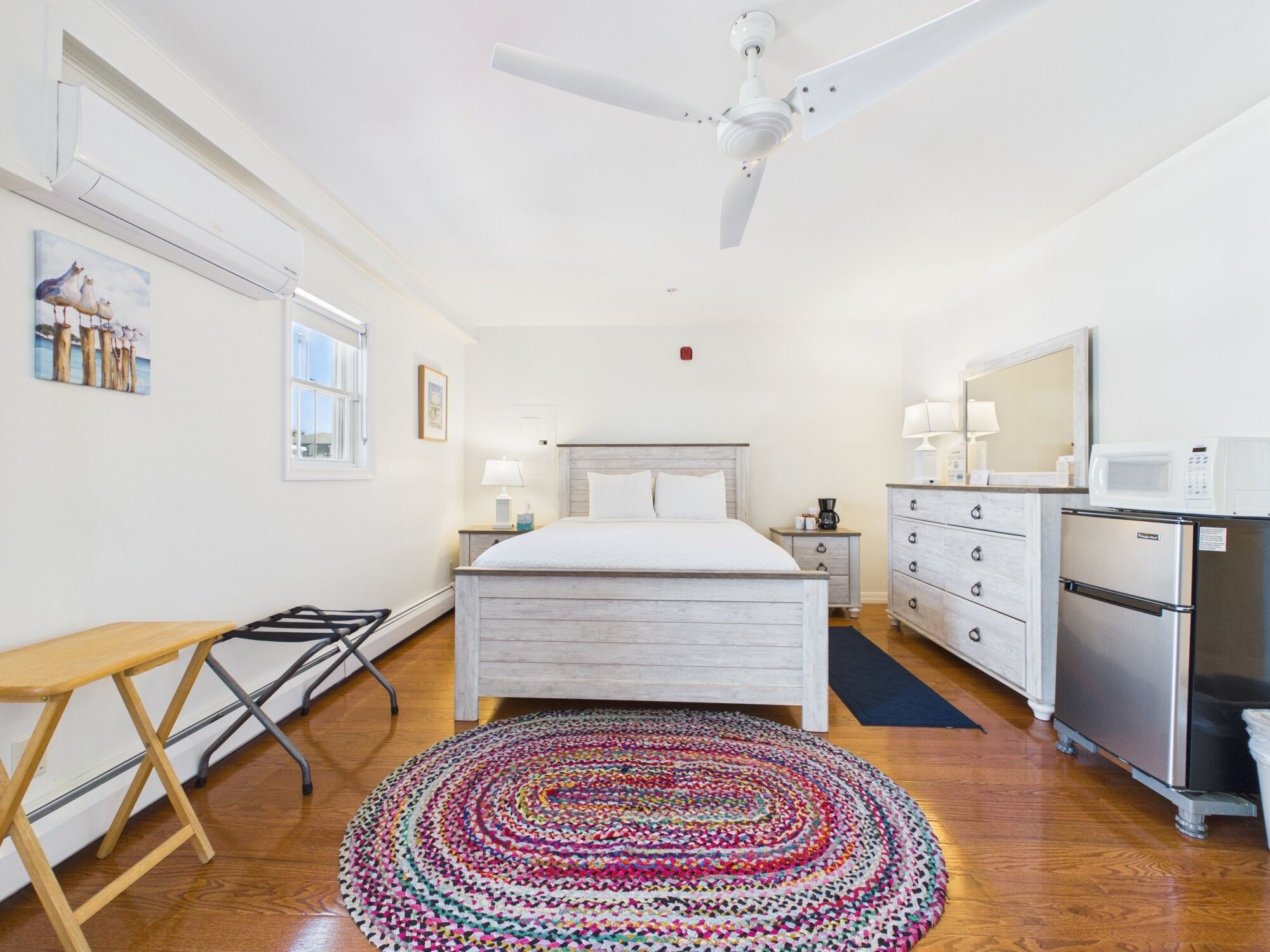 Un dormitorio con cama, mesitas de noche, escritorio, silla, ventilador de techo, microondas, refrigerador y una alfombra colorida.