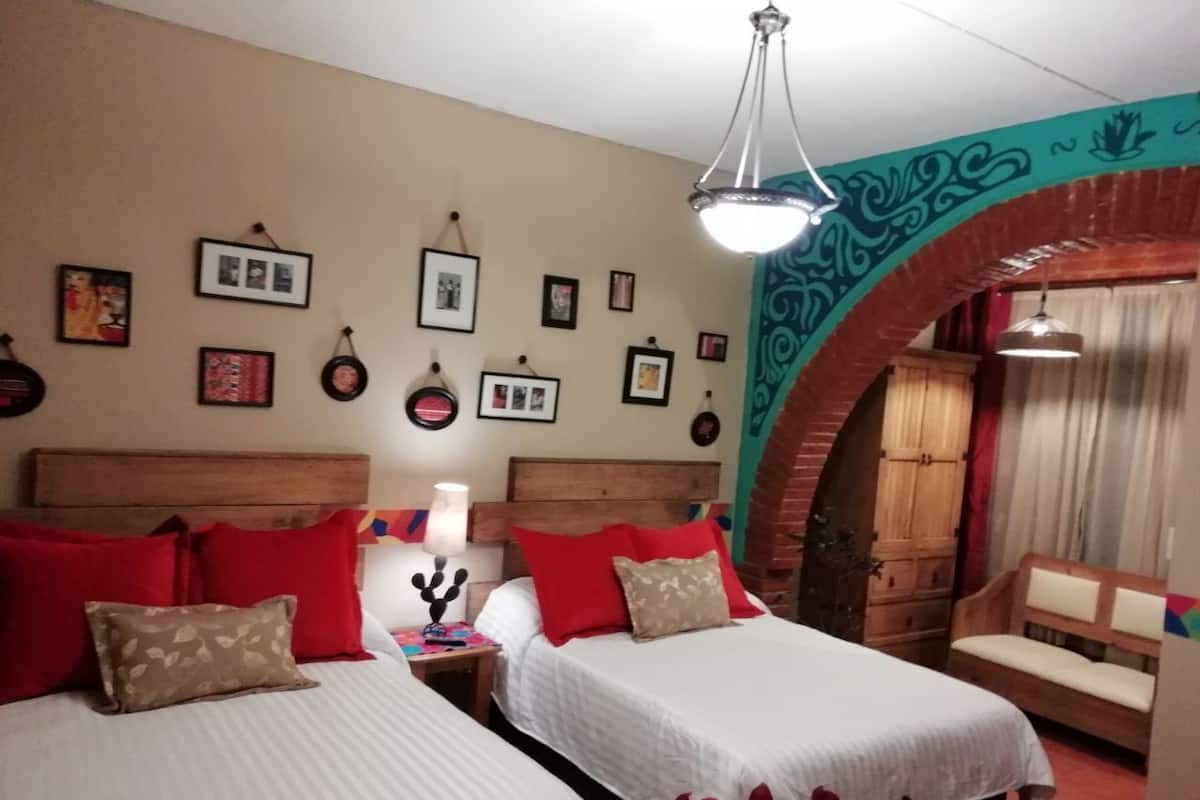 Habitación de hotel con dos camas, almohadas rojas y color beige, una mesita de noche con lámpara y un banco de madera.