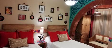 A hotel room with two beds, red and beige pillows, a nightstand with a lamp, and a wooden bench.