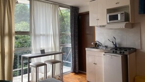 A compact room with a bed, a small table with two chairs, a kitchenette with a microwave and stove, and a large window with curtains.
