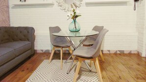 A dining area with a glass table and two chairs, a sofa, a decorative mirror, and shelves with plants and a phone.