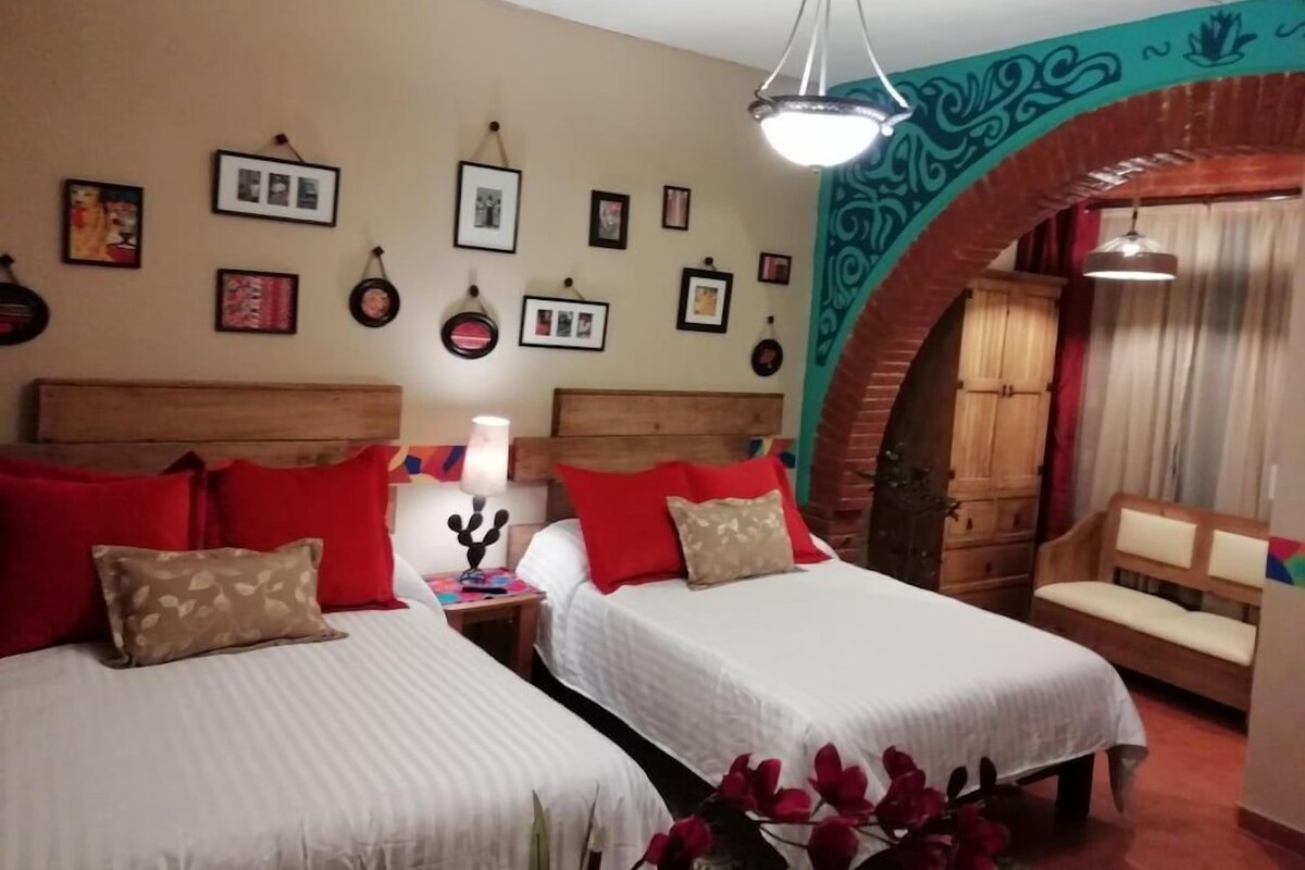 Ein Hotelzimmer mit zwei Betten, einem hölzernen Kopfteil, roten Kissen, einer Wandleuchte und einer dekorativen Rundbogenarchitektur.